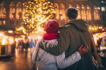 Couple facing toward a Christmas tree that is lit up. They have their arms wrapped around each others backs and are wrapped up warm.